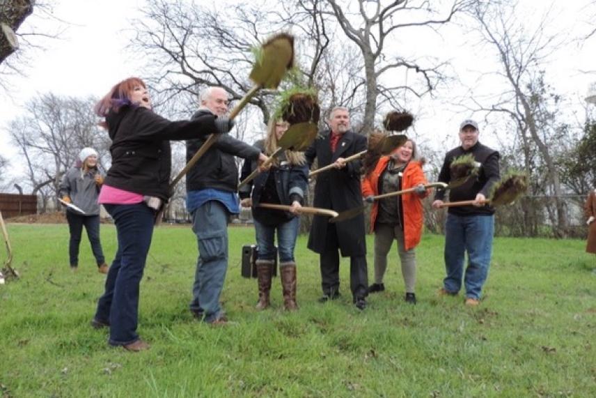 Seven people in a field holding shovels with soil, trees in the background.