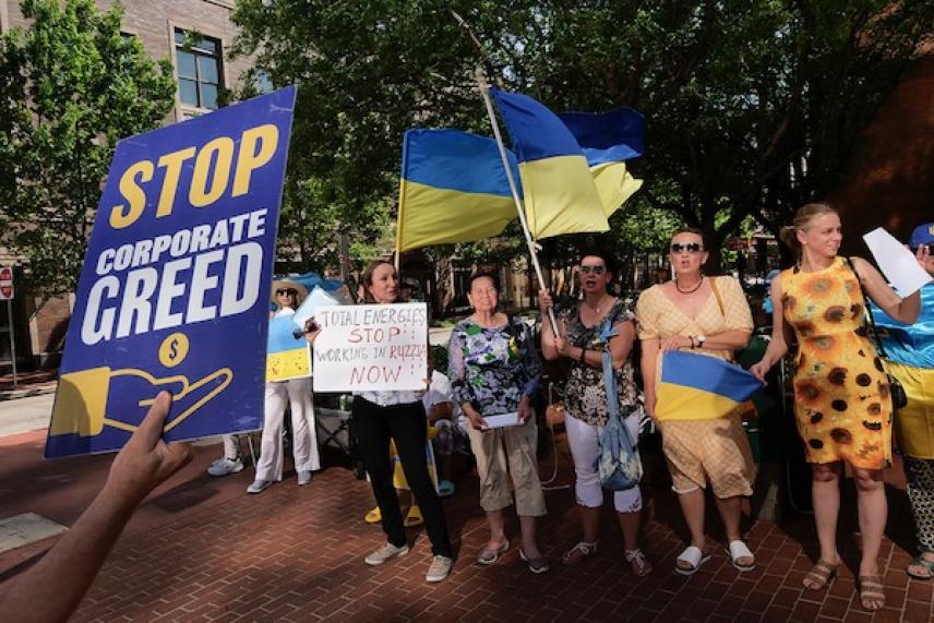 Protesters holding signs and Ukraine flags on a sunny street.