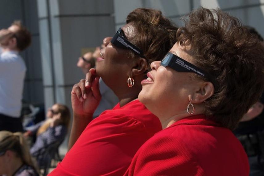 Two women in red shirts wearing eclipse glasses, looking up at the sky.