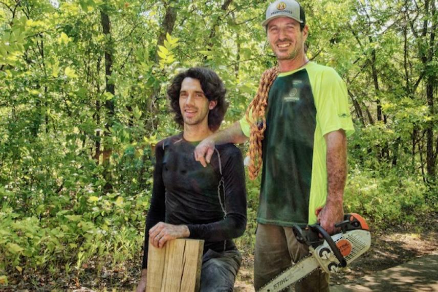 Two men in wooded area; one holding wood, the other a chainsaw, both smiling.