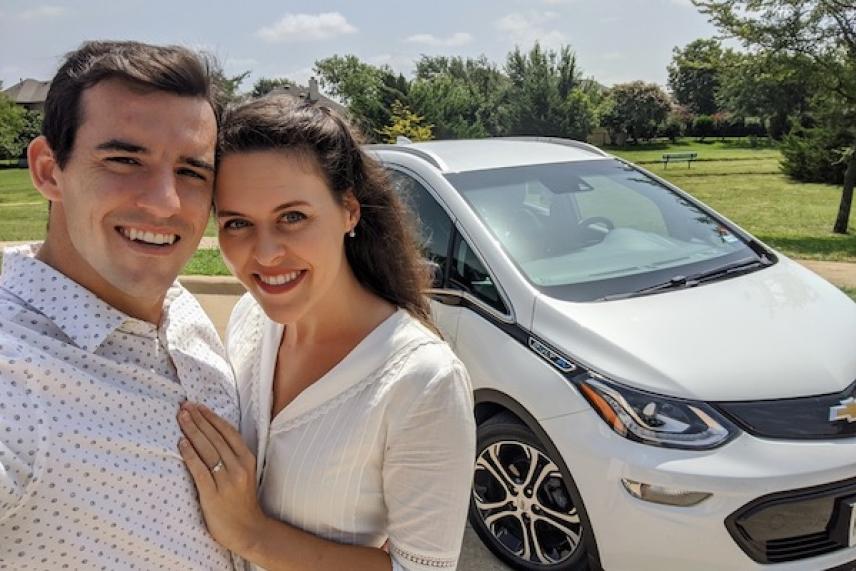 Couple smiling in front of a white car on a sunny day.