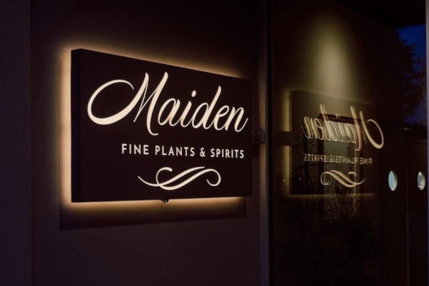 Illuminated sign reading "Maiden, Fine Plants & Spirits" with reflection on glass.