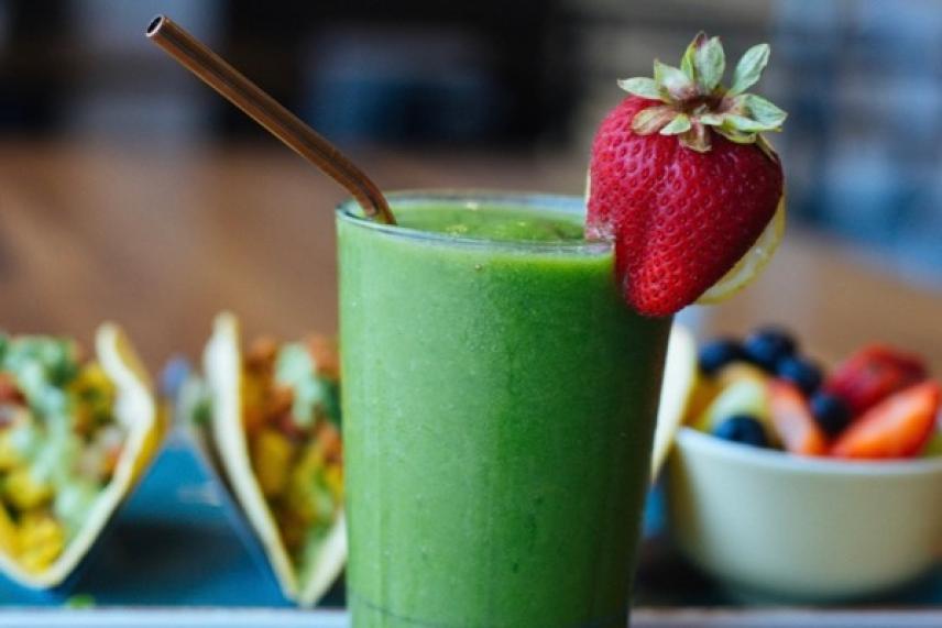 Green smoothie with a strawberry on the rim, tacos and fruit bowl in the background.
