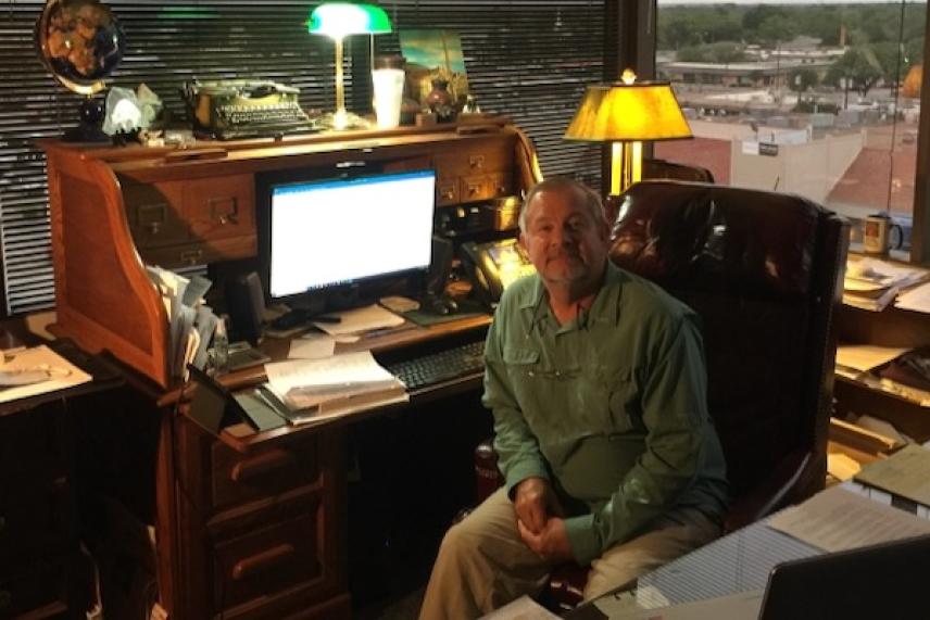 Man sitting at a wooden desk with a computer in a dimly lit office.