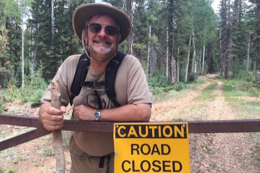 Smiling man in outdoor gear by "Caution Road Closed" sign, forest background.
