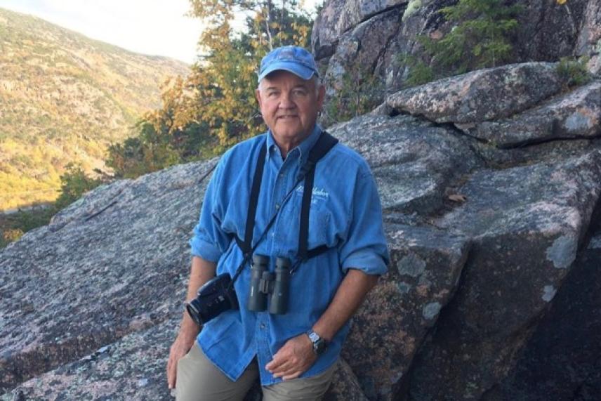 Man in blue shirt with binoculars, standing on rocky terrain.
