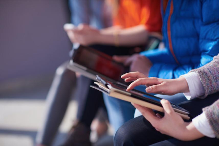 Hands using tablets, people seated, wearing colorful casual clothes.