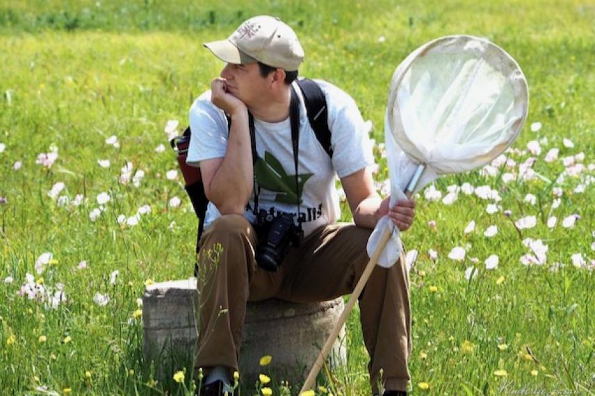 Person sitting on a log in a field, holding a butterfly net, wearing a hat and camera around neck.
