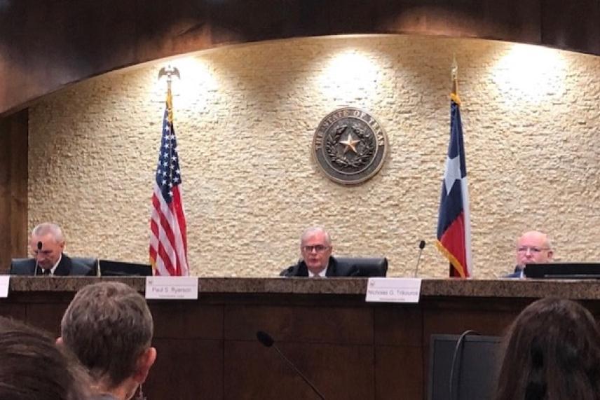 Three officials seated at a panel; US and Texas flags behind them.