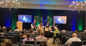 Panel discussion in a conference room with two screens and chandeliers overhead.