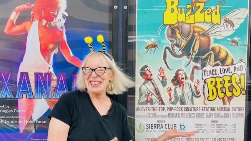 Smiling woman with antennae headband gestures at bee-themed poster.