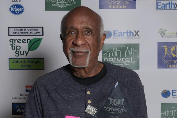 Elderly man smiling at a formal event, standing against a backdrop with logos.