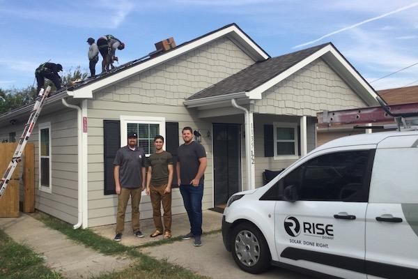 Workers installing solar panels on a house roof; a van with a logo is parked nearby.