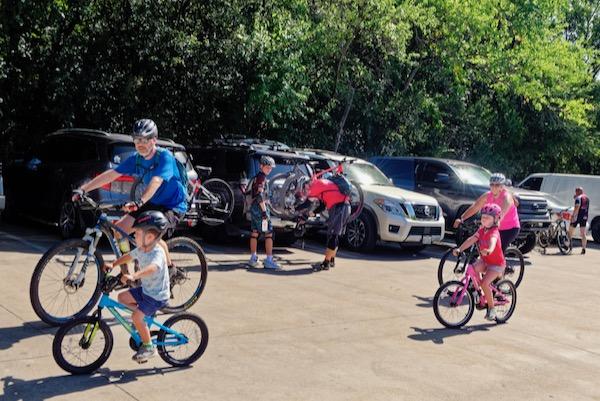 Families preparing bicycles in a sunny parking lot near greenery.