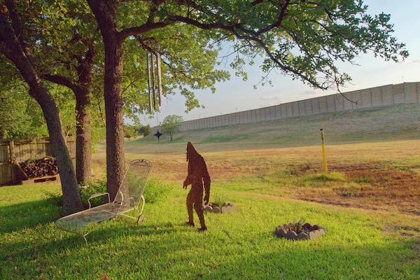 A person in a brown costume stands under a tree with a wind chime on a grassy field.