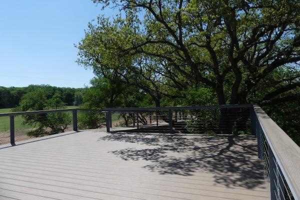 Wooden deck with metal railing, shaded by leafy trees, under a clear blue sky.