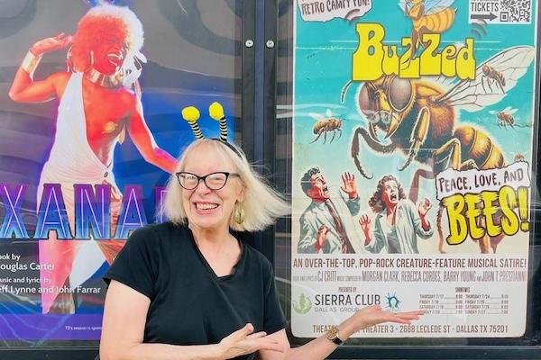Smiling woman with antennae headband gestures at bee-themed poster.