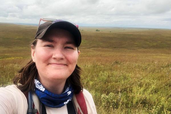 Smiling person in a cap and scarf standing in a vast grassy field.
