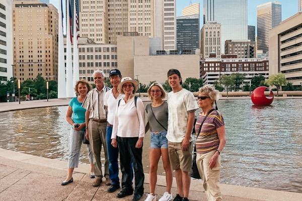 Group of seven people smiling in front of a city fountain and skyscrapers.
