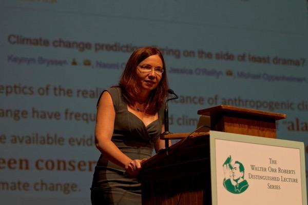 A woman speaks at a podium with a presentation slide in the background.