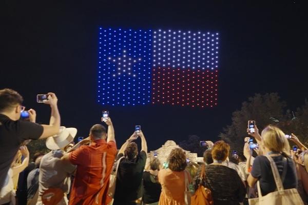 Crowd watches Texas flag light display at night, taking photos.