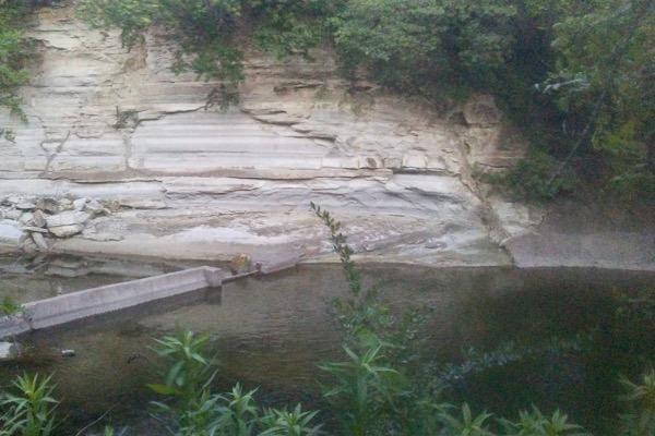 Rocky stream with green foliage, weathered cliff, and a small wooden bridge.