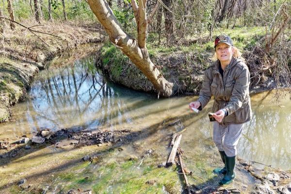 Woman in a cap stands by a creek, holding a small object, surrounded by trees.