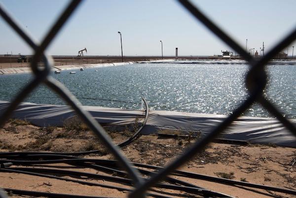 Chain-link fence view of a sparkling water reservoir and distant oil pumps.