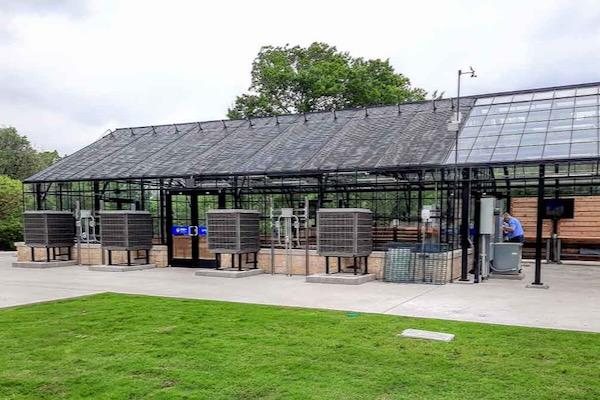 Glass greenhouse with large air conditioning units outside, set on a concrete path and green grass.