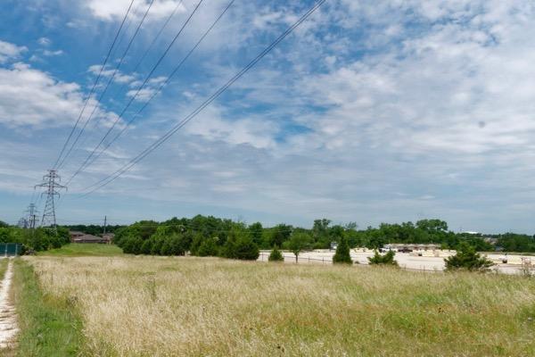 Power lines over a grassy field and trees under a partly cloudy sky.