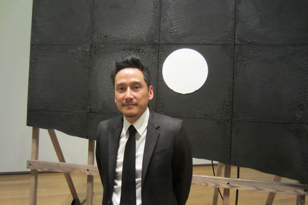 Man in a suit stands before a black artwork with a white circle.