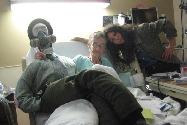 Hospital room with three people; one wearing a gas mask, others smiling.