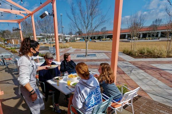 Server with a mask serves food to a group seated outdoors under a pergola.