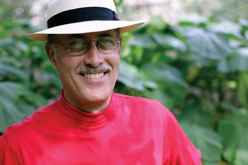 Smiling man in a white hat and red shirt, outdoors with green leaves in the background.