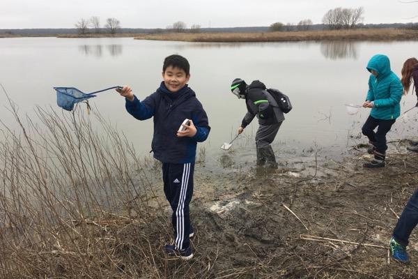 Children exploring a lakeshore with nets on a cloudy day.