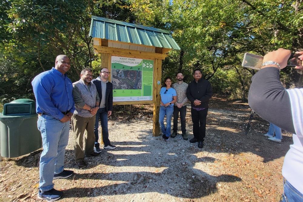 Group of people posing by a park information board, trees in the background.