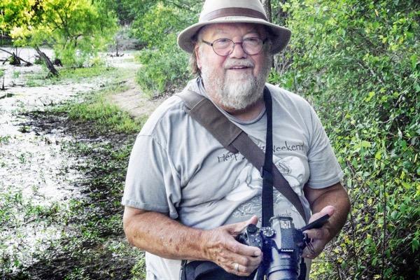 Elderly man with a camera and hat smiles in a lush, green outdoor setting.