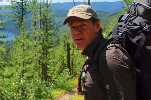Man hiking on a forest trail, wearing a backpack and cap, with mountains in the background.