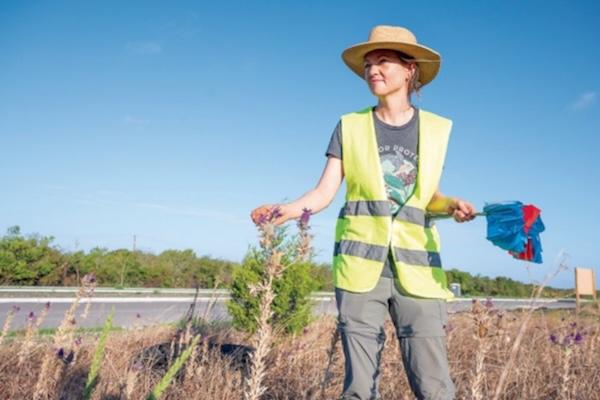 Worker in safety vest, straw hat holding flags, standing by a highway, clear sky.