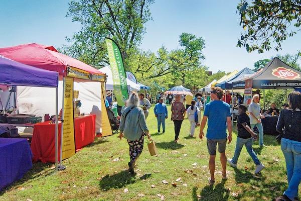 People walking between colorful market stalls on a sunny day in a park.