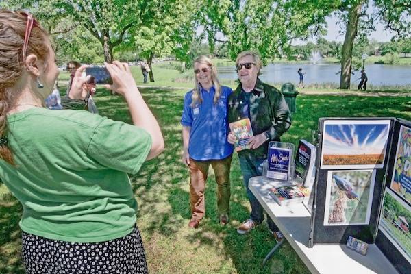 Person photographing two people at an outdoor event near a lake.
