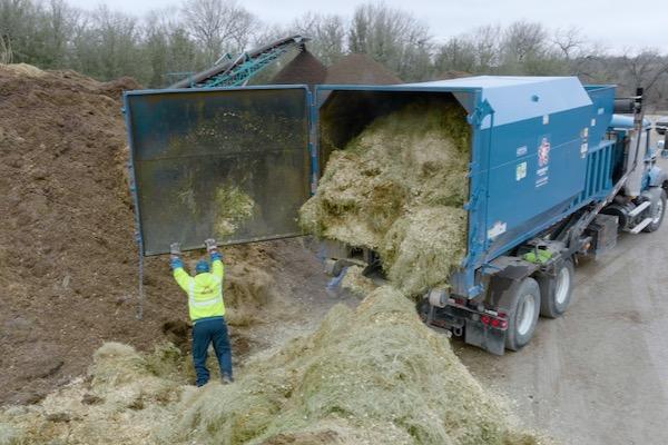 Truck offloading hay into a large pile, person in yellow vest nearby.