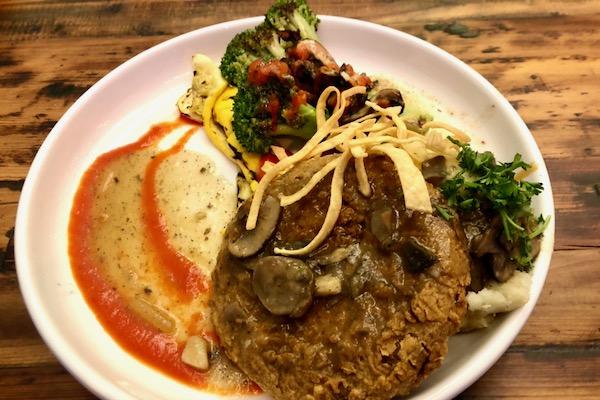 Plate of vegetables, mashed potatoes, and patty garnished with sauce and herbs.