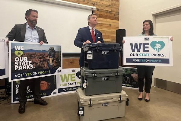 Three people hold signs promoting state parks inside a room with stacked coolers.