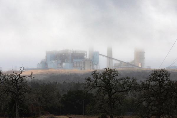 Factory obscured by mist, surrounded by trees, under a cloudy sky.