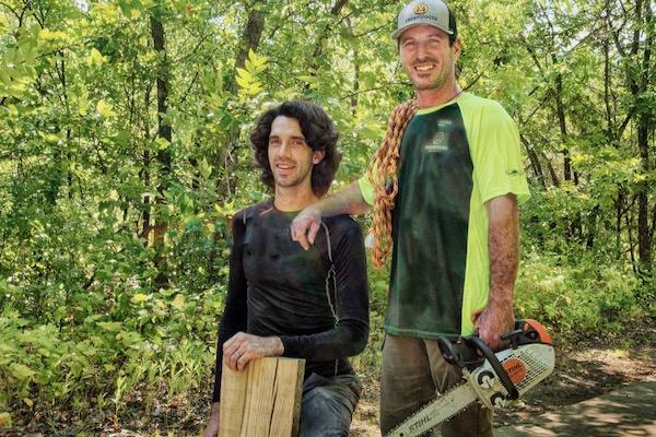 Two men in wooded area; one holding wood, the other a chainsaw, both smiling.