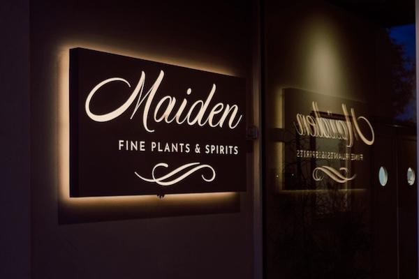 Illuminated sign reading "Maiden, Fine Plants & Spirits" with reflection on glass.