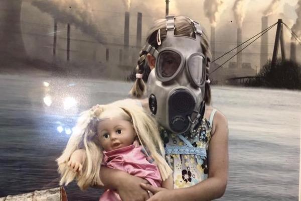 Child wearing gas mask, holding doll in polluted industrial landscape.