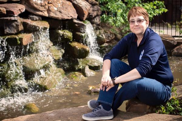 Seated person by a small waterfall in a garden setting, wearing a navy shirt and jeans.