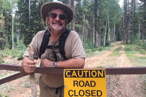 Smiling man in outdoor gear by "Caution Road Closed" sign, forest background.